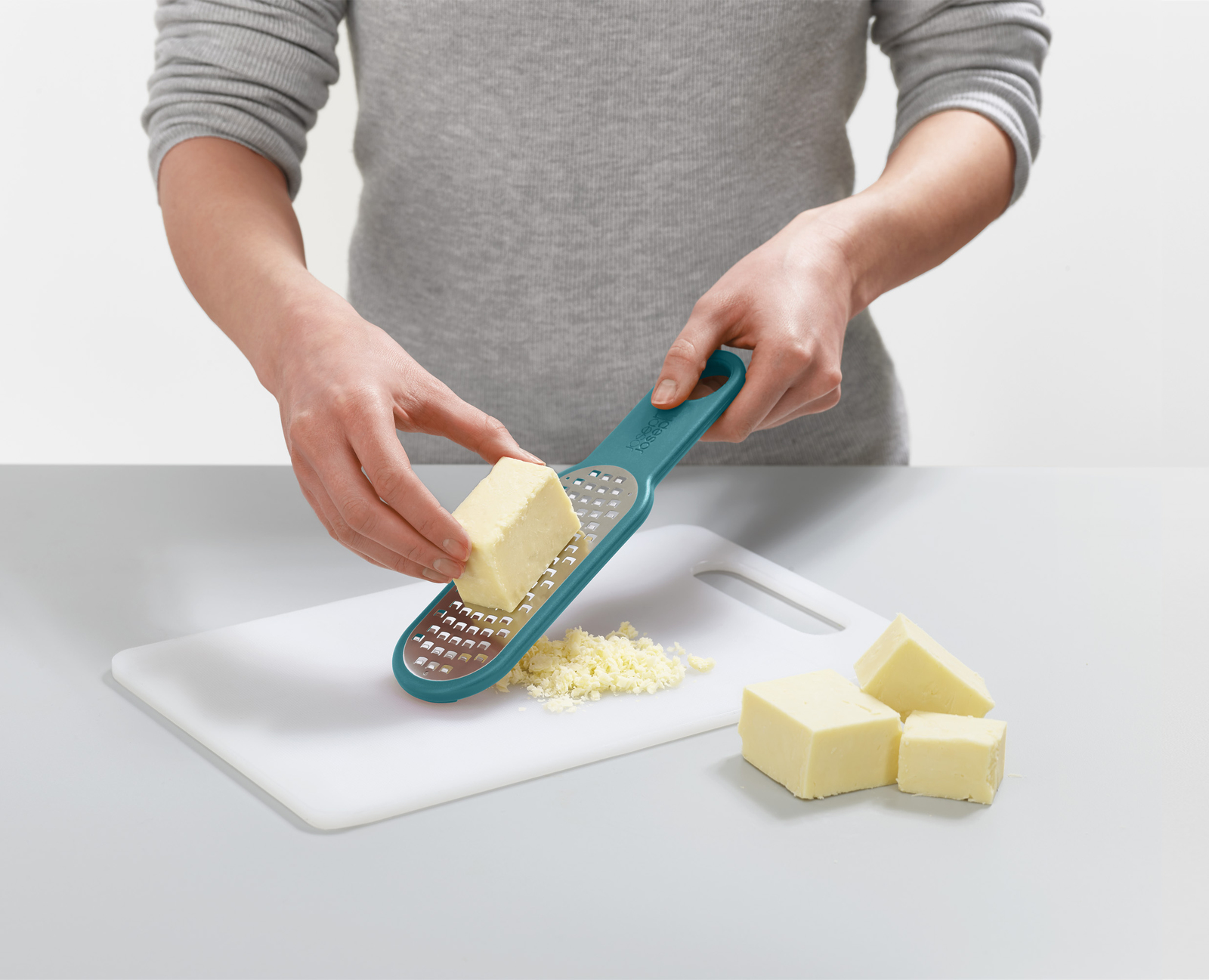 Una persona grattugia il formaggio su un tagliere bianco. Sono presenti blocchi di formaggio e formaggio grattugiato, con una grattugia turchese.