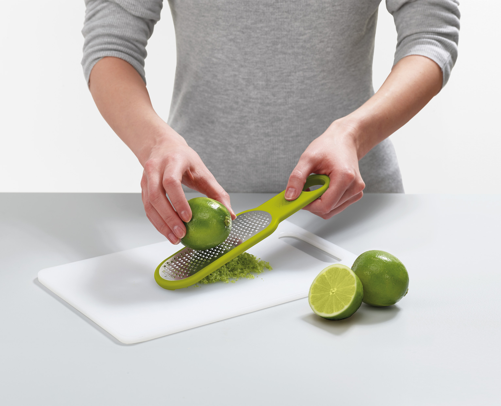 Una persona grattugia un lime con una grattugia verde su un tagliere bianco. Due lime a destra.
