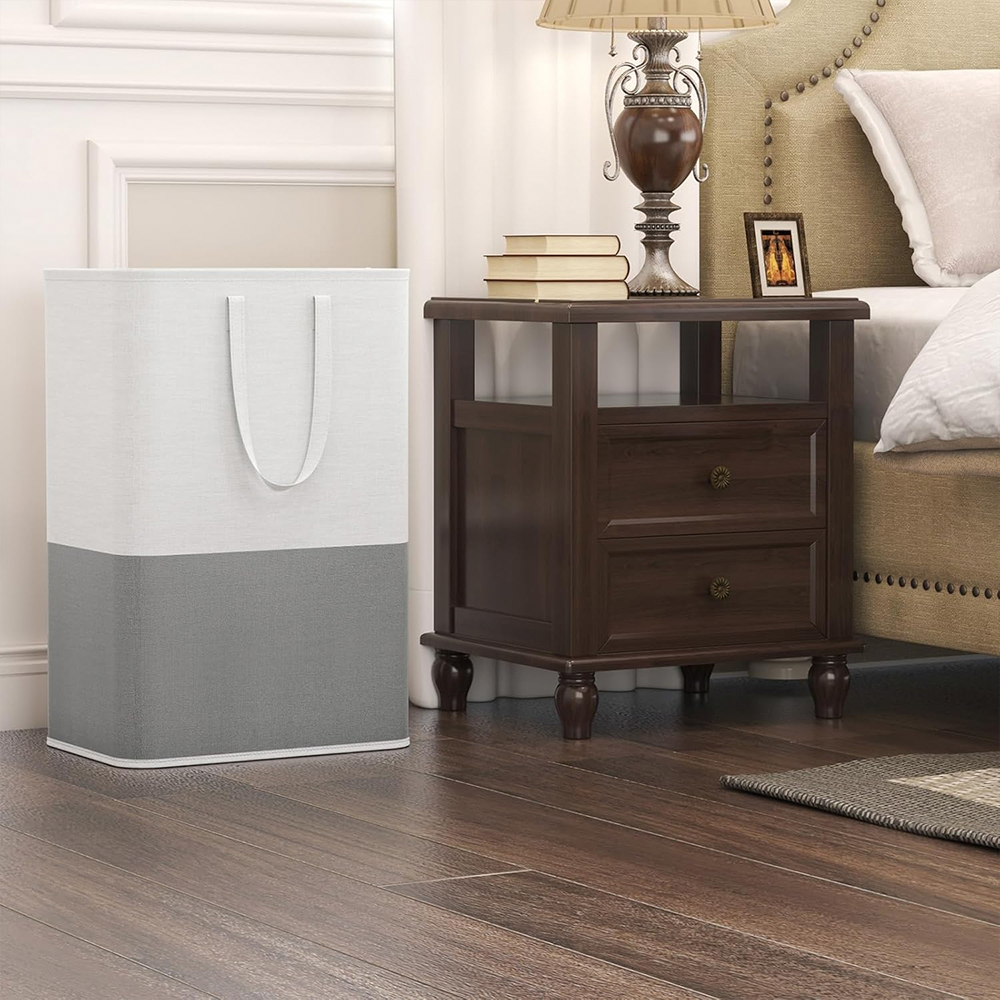Escena de dormitorio con cesto de ropa sucia, mesita de noche, cama y suelo de madera. El cesto es blanco y gris. La mesita es de madera oscura.