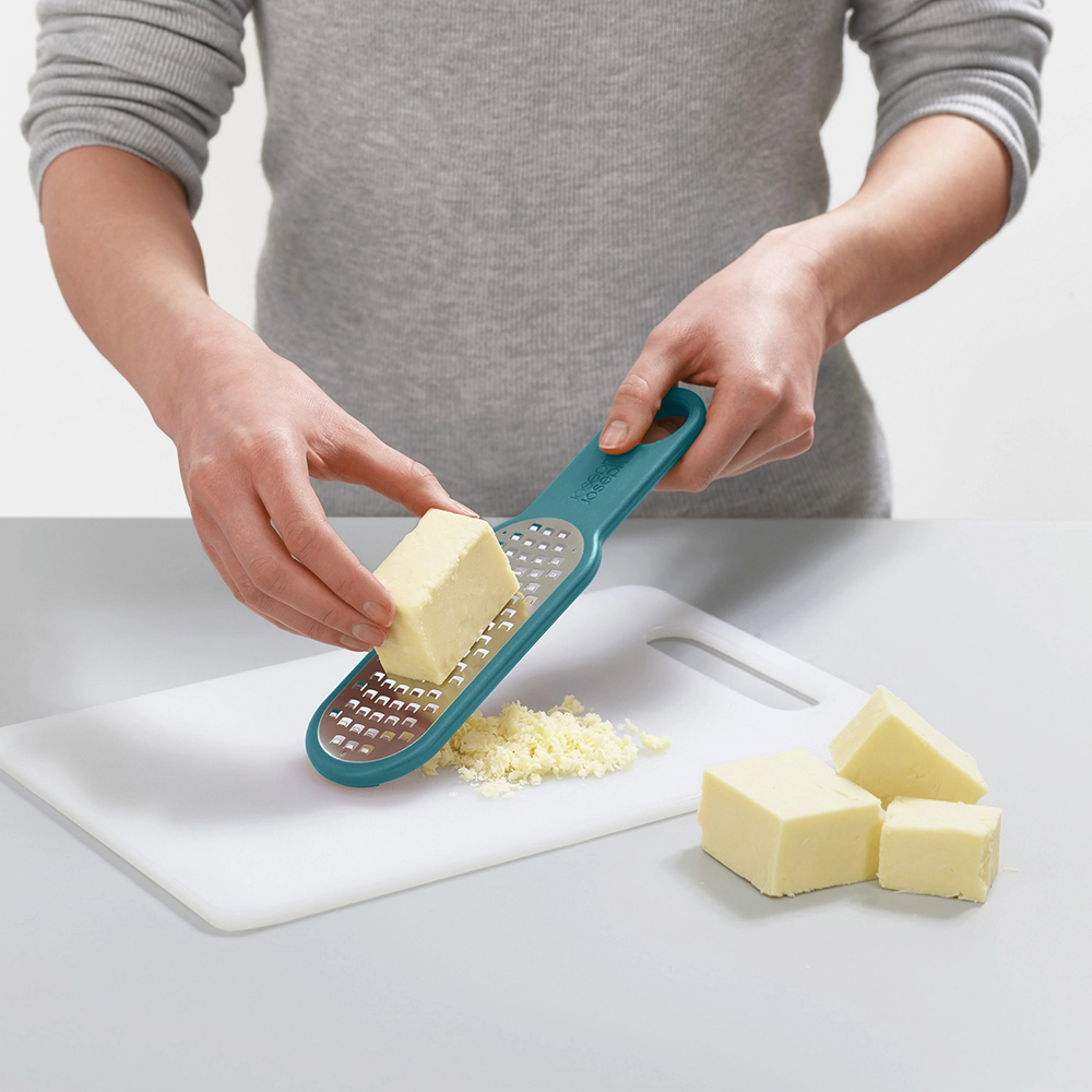 Una persona grattugia il formaggio con una grattugia turchese su un tagliere bianco. Si vedono formaggio grattugiato e blocchi di formaggio.