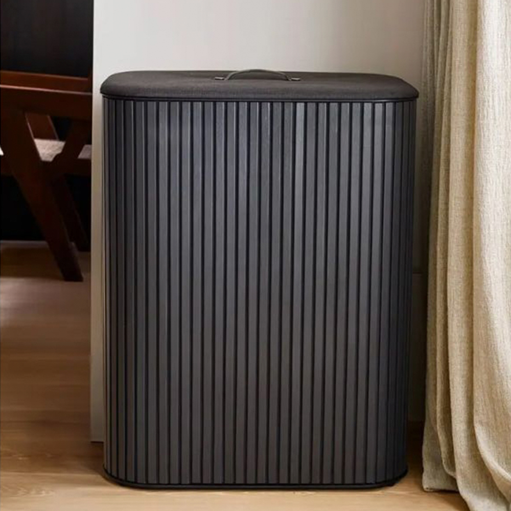 Un cesto de ropa sucia gris oscuro y acanalado con tapa plana y asa. Una silla de madera y una cortina están en el fondo.