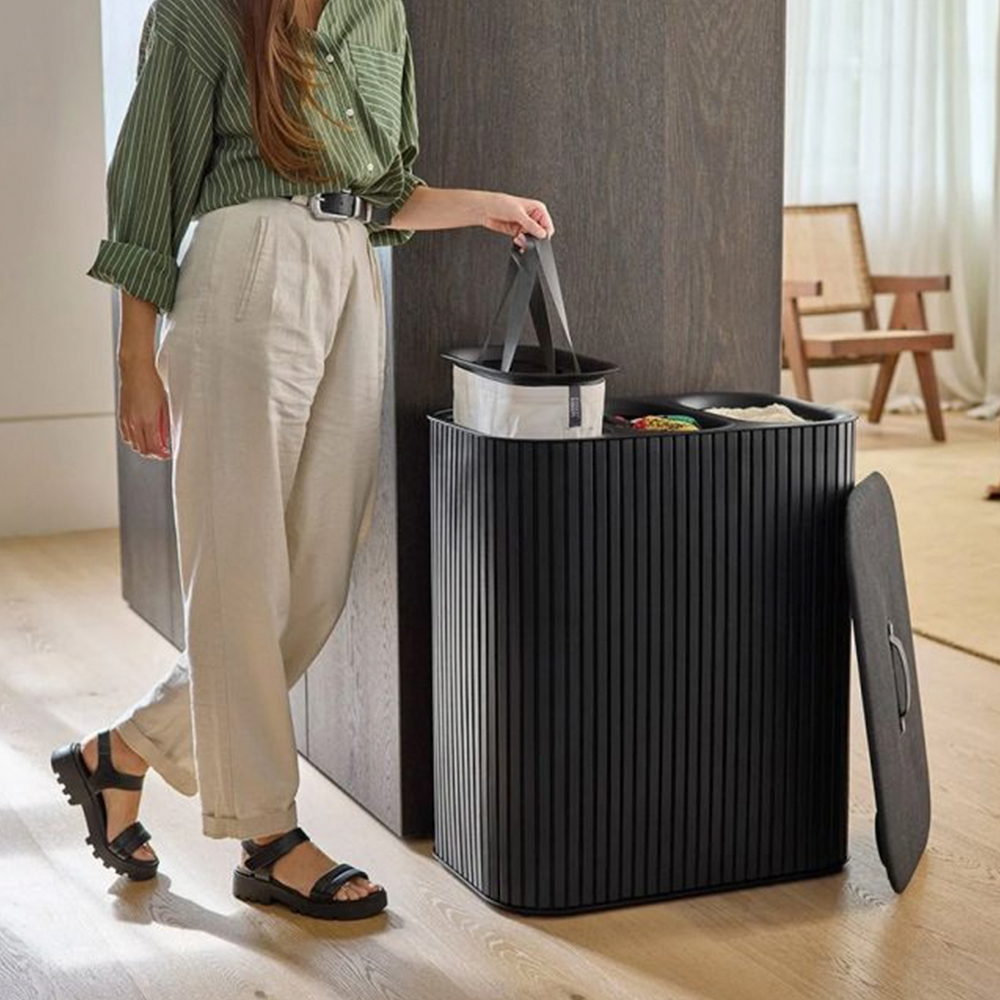 Una mujer está de pie cerca de un cubo negro, sosteniendo una bolsa. El cubo tiene una tapa abierta. La mujer lleva una camisa verde y pantalones beis.