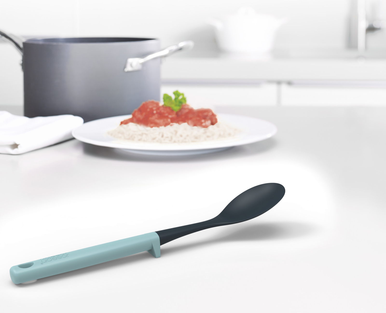 Una escena de cocina con una cuchara en foco. Plato con comida en el fondo. Olla y otros elementos de cocina son visibles.