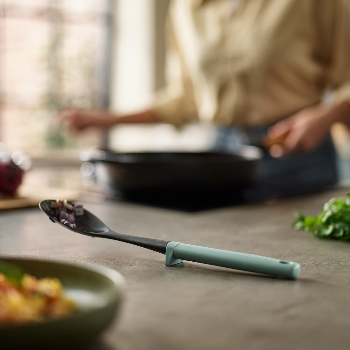 Escena de cocina con una cuchara de cocina que contiene cebollas picadas. Una persona cocina en el fondo. Un cuenco verde y hierbas son visibles.