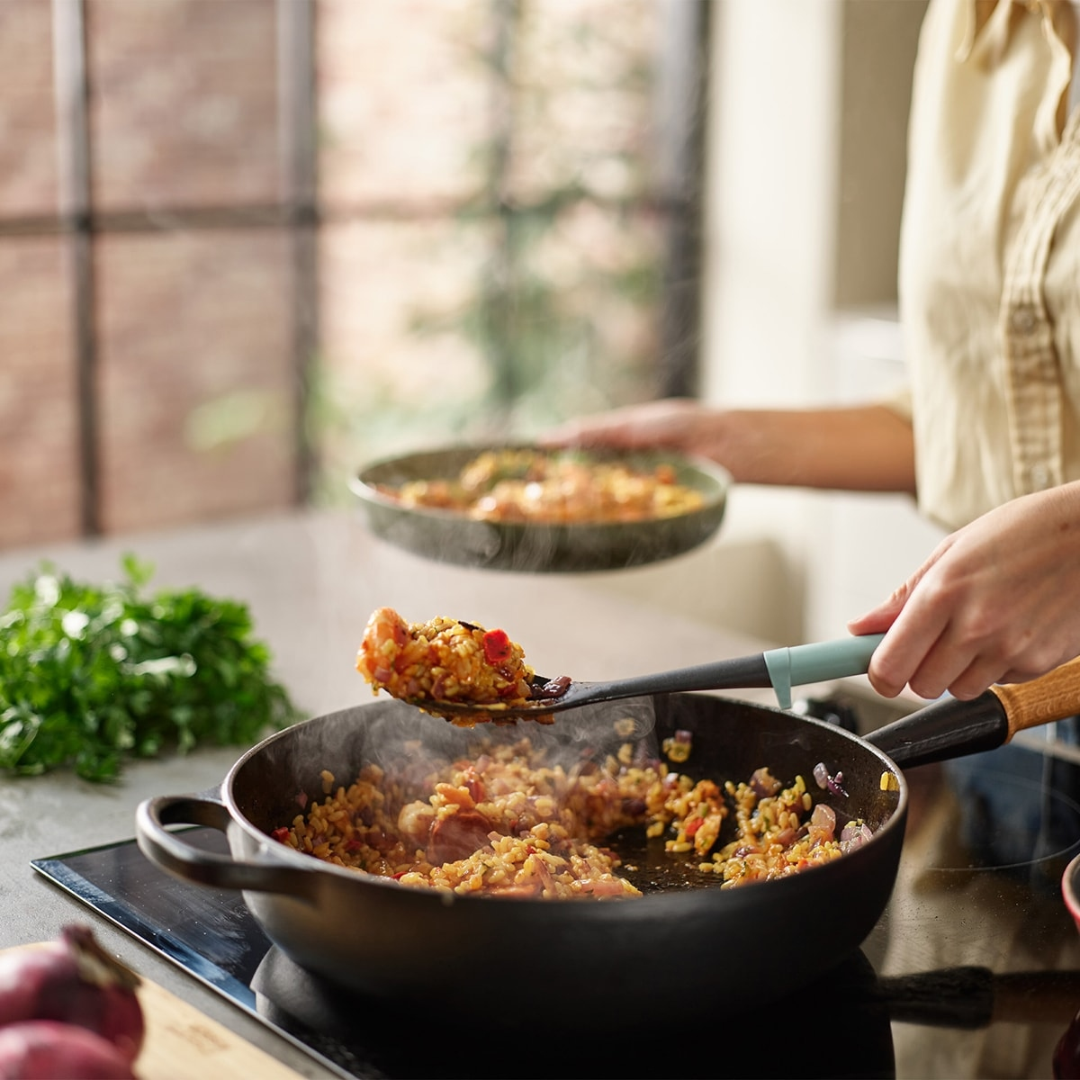 Una persona cocina en una sartén. Sale vapor de la comida. Una cuchara saca comida de la sartén a un plato. Perejil y cebollas están en la encimera.