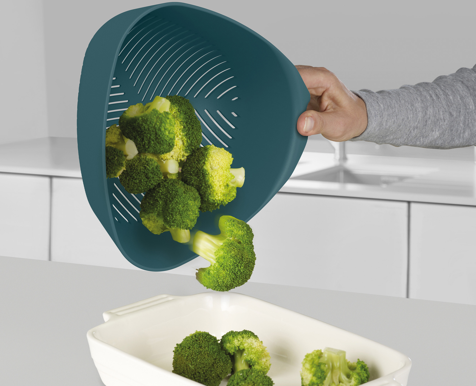Mano vertiendo brócoli de un colador verde azulado a una fuente blanca. Fondo de cocina.