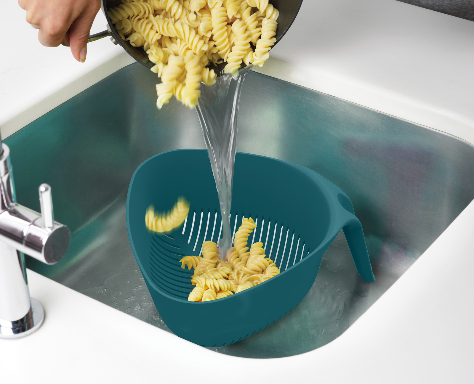 La pasta se vierte de una sartén a un colador verde azulado en un fregadero. El agua fluye sobre la pasta.