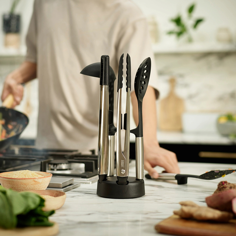 Utensilios de cocina en un soporte sobre una encimera. Una persona está cocinando al fondo. Verduras y especias en la encimera.