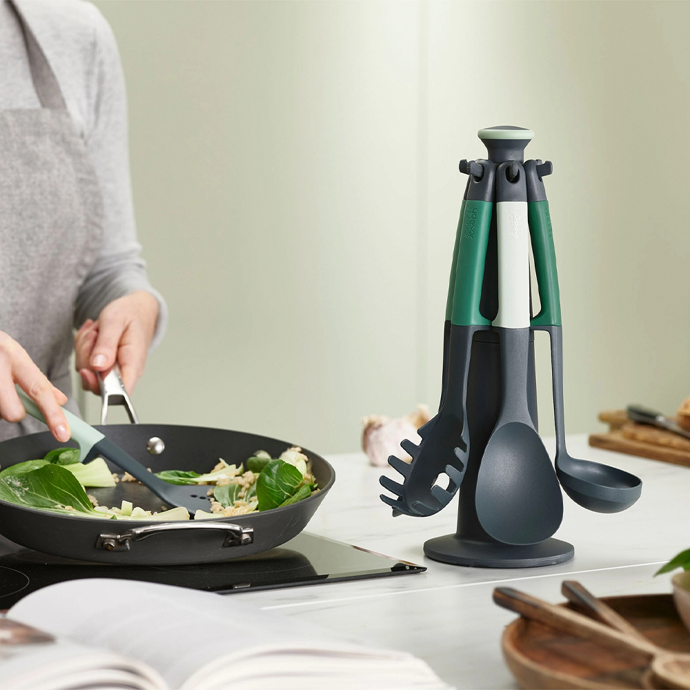 Una persona cocina en una sartén, utensilios de cocina en un soporte. Se están cocinando verduras. Un libro de cocina está abierto.