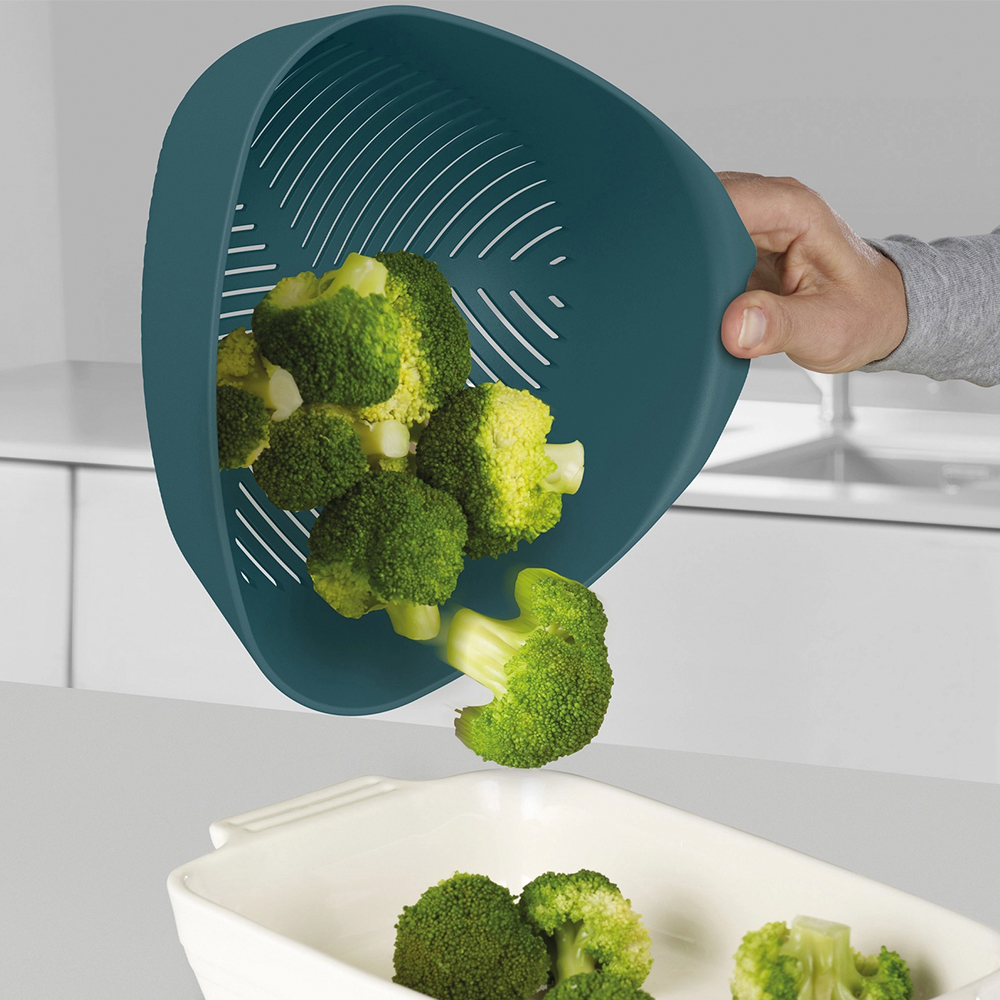 Una persona vierte brócoli de un colador verde azulado a un plato blanco. Los ramilletes de brócoli están en el colador y el plato.