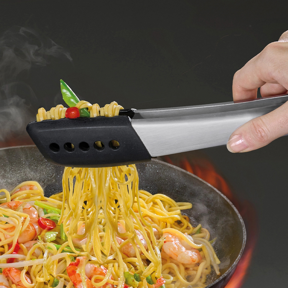 Pinze che tengono spaghetti con verdure e un pomodoro. Gli spaghetti cadono in una padella con gamberi e verdure.