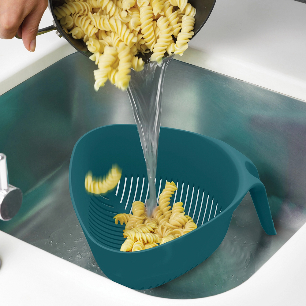 La pasta se vierte de una olla a un colador verde azulado en un fregadero de acero inoxidable. El agua sale a chorros de la olla, enjuagando la pasta.