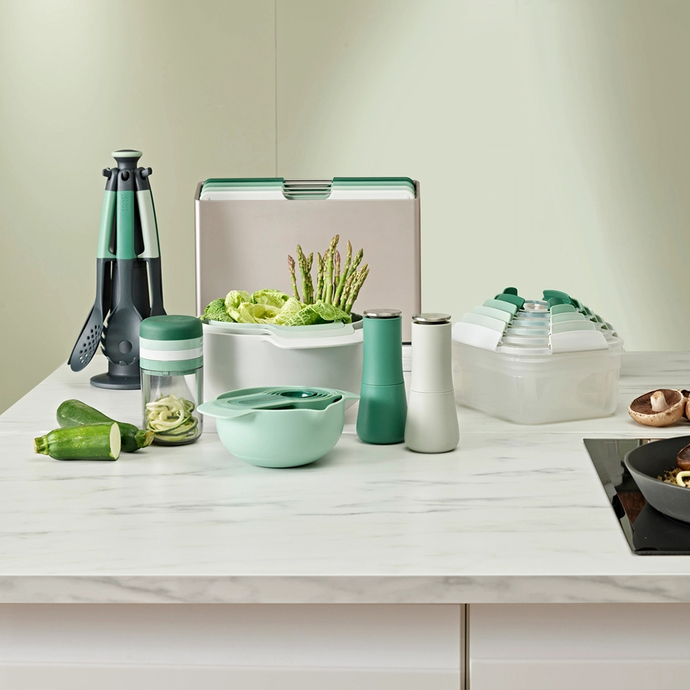 Utensilios de cocina y verduras en una encimera. Incluye tabla de cortar, recipientes, calabacín, espárragos y tazas medidoras.