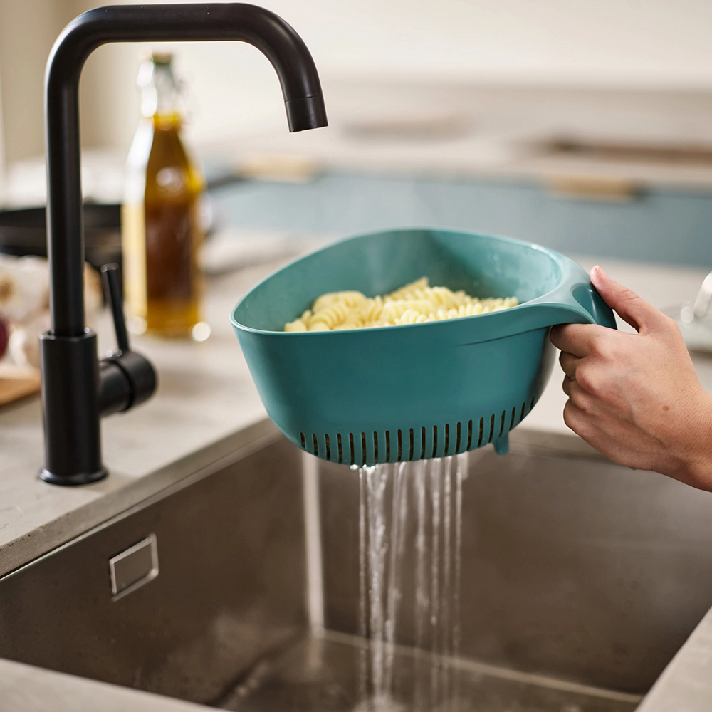 Una persona enjuaga pasta en un colador de color verde azulado sobre un fregadero de cocina. Un grifo negro está arriba. Botella de aceite de oliva en el fondo.