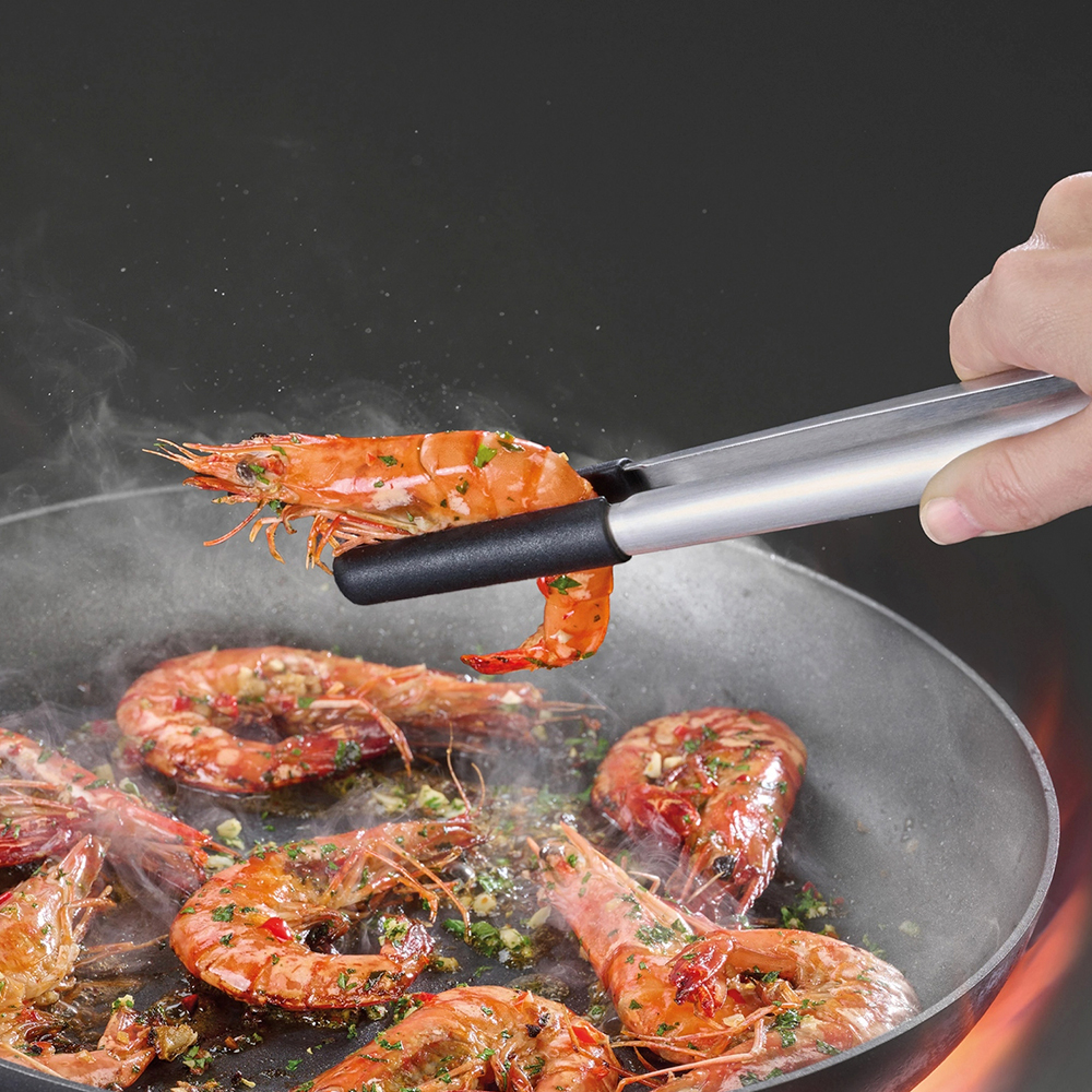 Una mano tiene delle pinze che sollevano un gambero cotto da una padella con altri gamberi. Il vapore sale. La padella è sopra le fiamme.