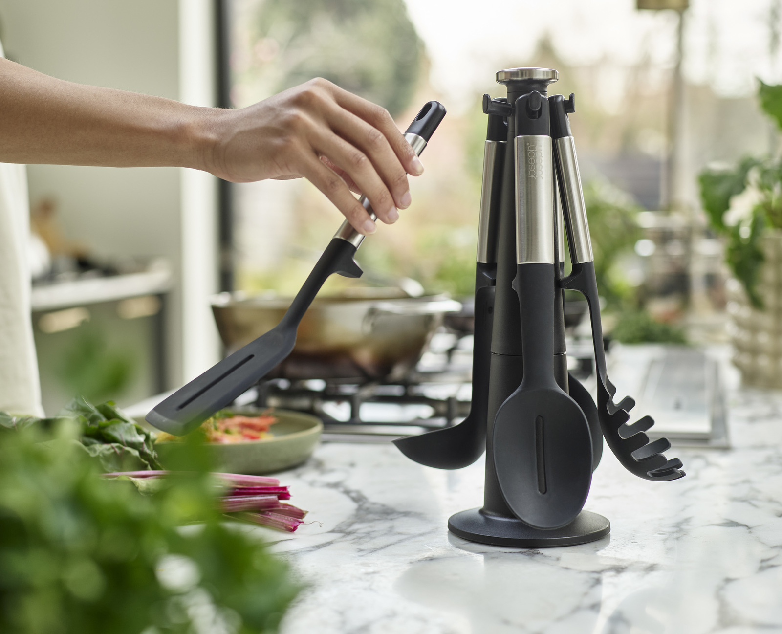 Una mano sostiene una espátula negra de un juego de utensilios de cocina. Otros utensilios están en un soporte negro. Verduras y una olla en el fondo.
