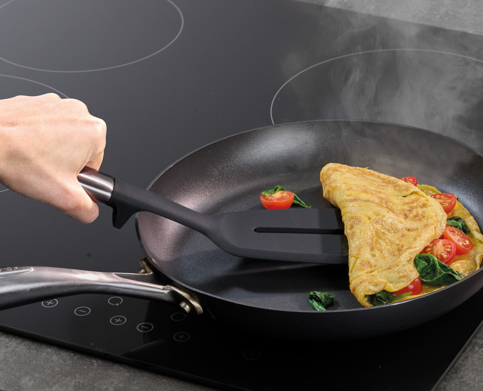 Una mano voltea una tortilla en una sartén con una espátula. La tortilla tiene tomates y espinacas. Sale vapor de la sartén.