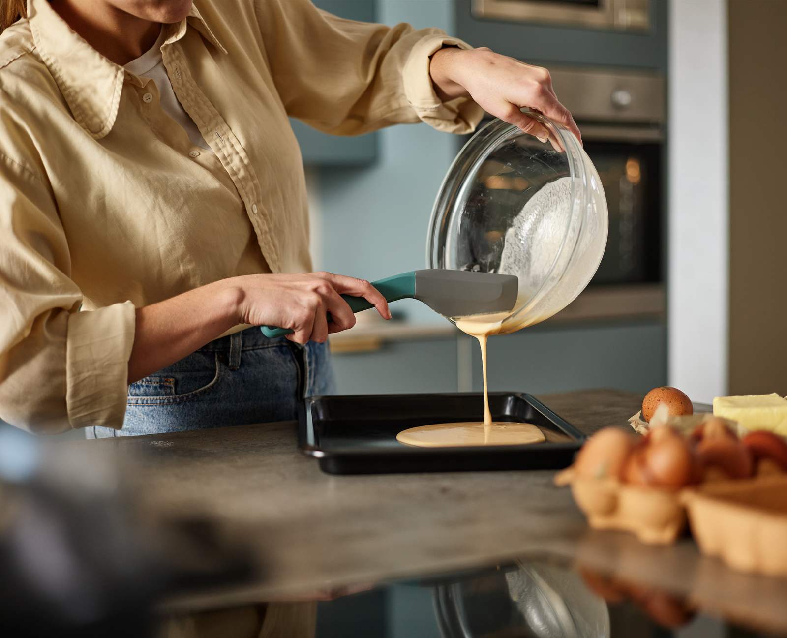 Una persona vierte la masa de un bol a una bandeja de horno. Hay huevos y mantequilla cerca. La persona lleva una camisa color canela y vaqueros.