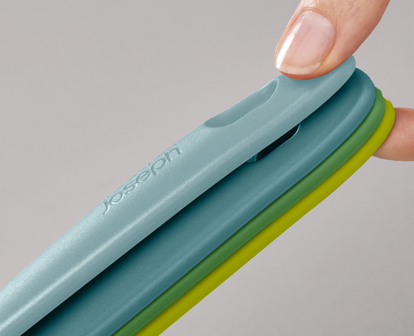 Una mano sostiene tres tablas de cortar de colores: azul, verde y amarillo. Las tablas están apiladas, y la superior se levanta.