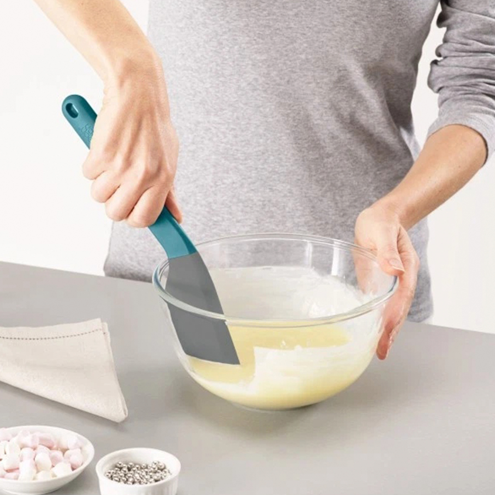Persona mezclando ingredientes en un bol de cristal con una espátula turquesa. Varios cuencos pequeños con aderezos y una servilleta están sobre la mesa.