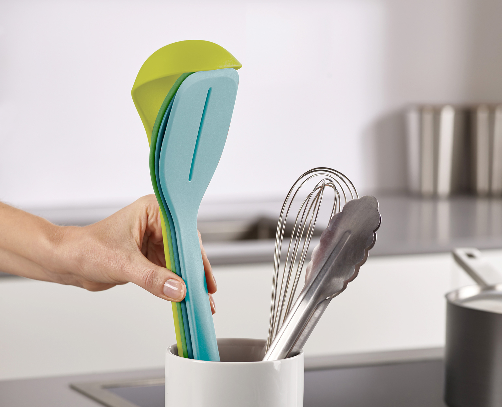 Una mano sostiene espátulas verdes, azules y turquesas en un recipiente blanco. Al lado hay un batidor y unas pinzas. El fondo tiene una cocina.
