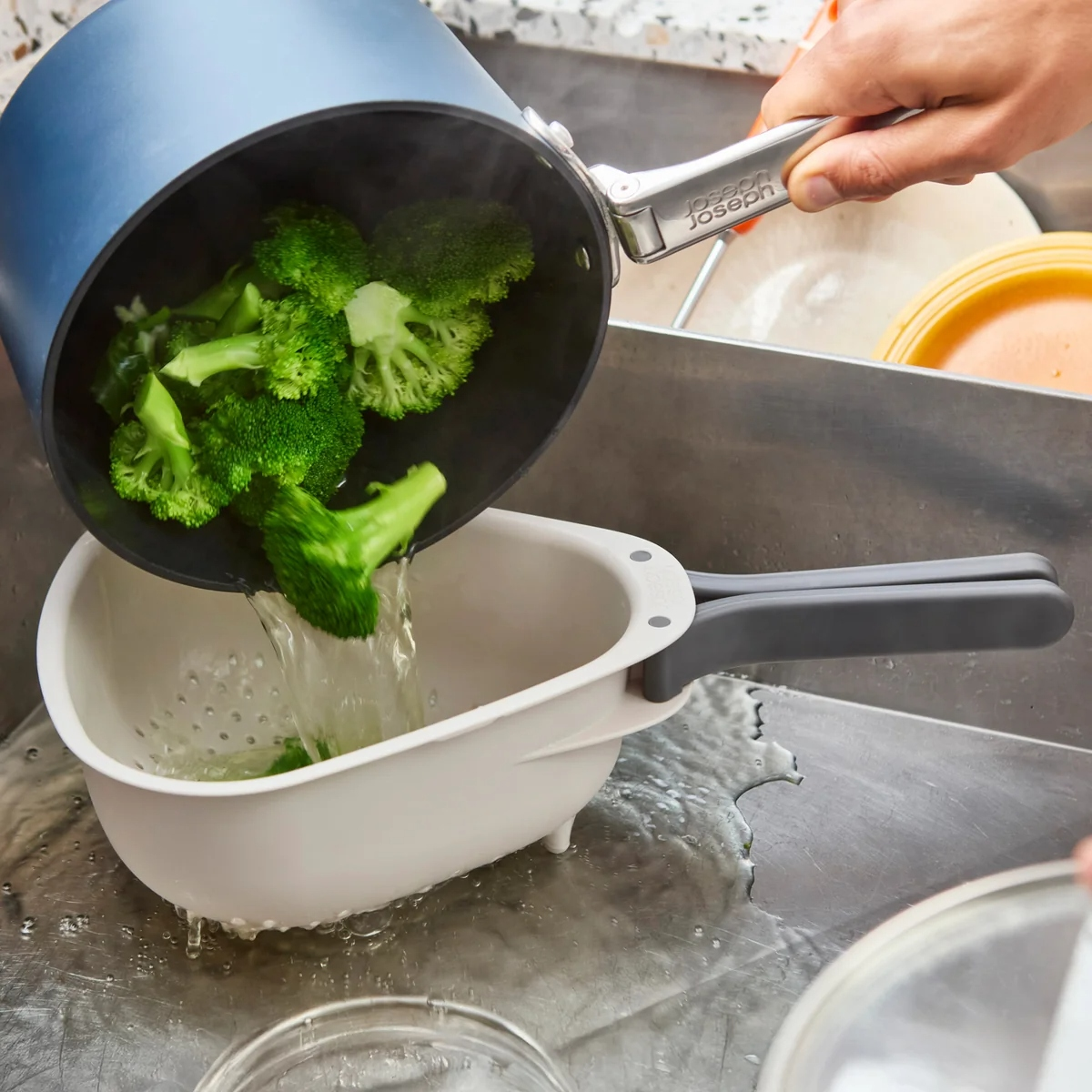 Una persona versa broccoli al vapore da una pentola blu in uno scolapasta bianco in un lavandino in acciaio inox. L'acqua scorre.