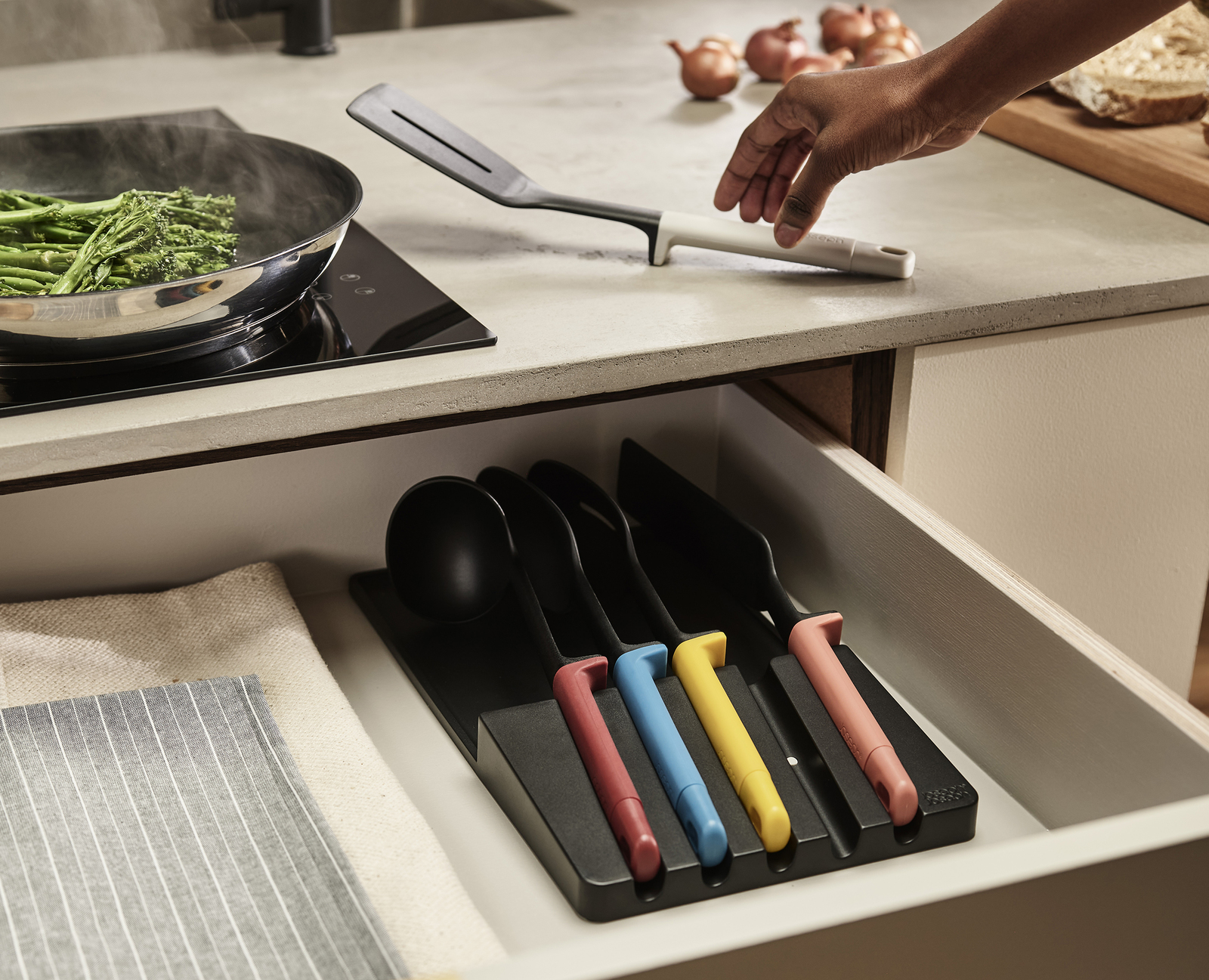 Una escena de cocina con una sartén de verduras, una mano que alcanza una espátula y utensilios coloridos en un cajón.