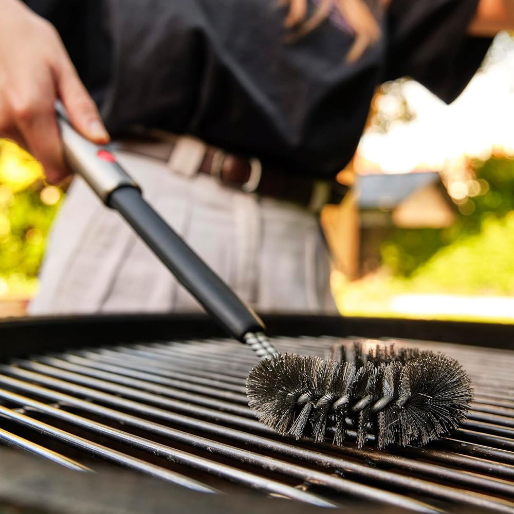 Una persona limpia una parrilla con un cepillo. El cepillo tiene cerdas metálicas, la parrilla es negra y el fondo está borroso.