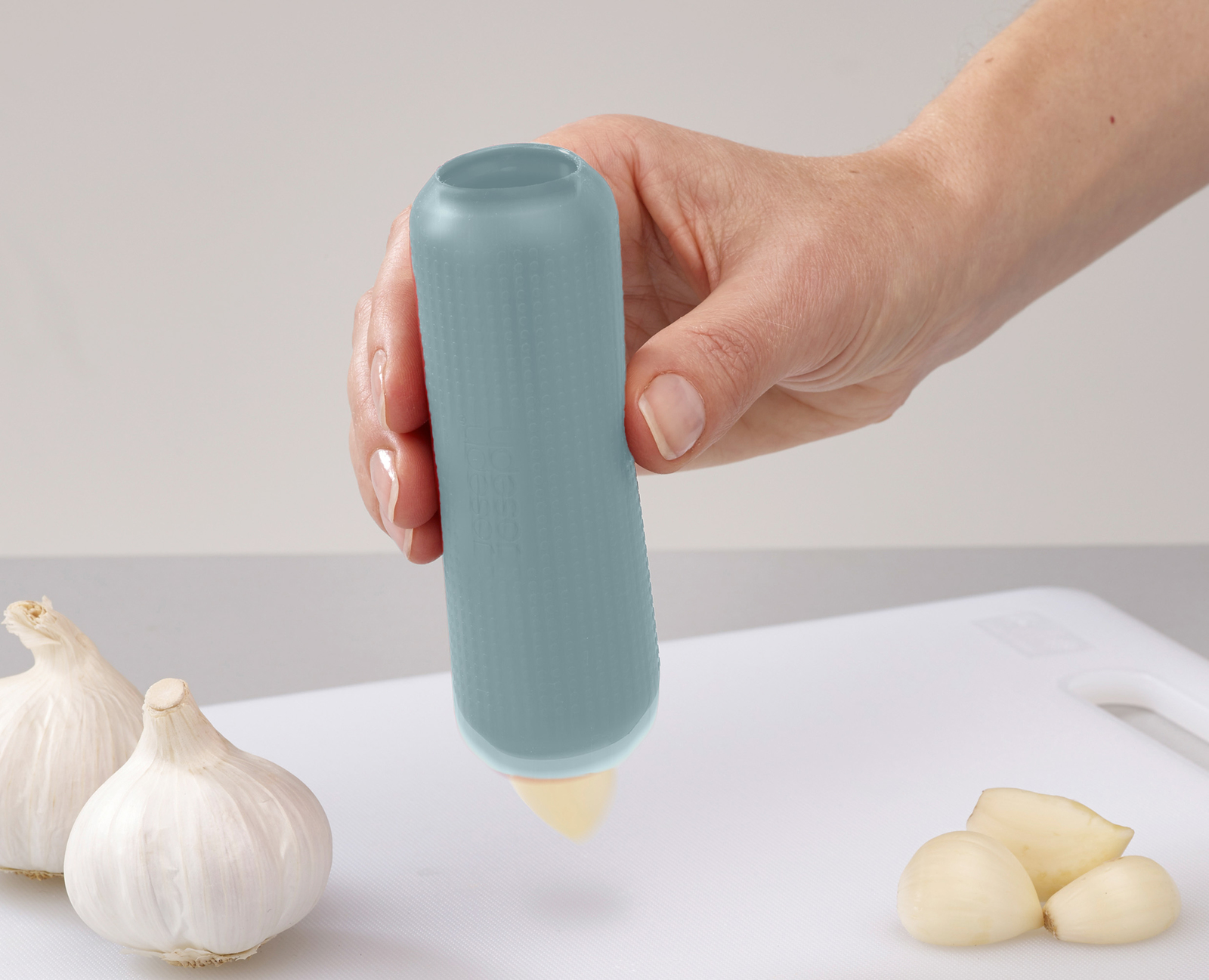 Una mano sostiene un prensador de ajo azul sobre una tabla de cortar blanca. Hay dientes y bulbos de ajo.