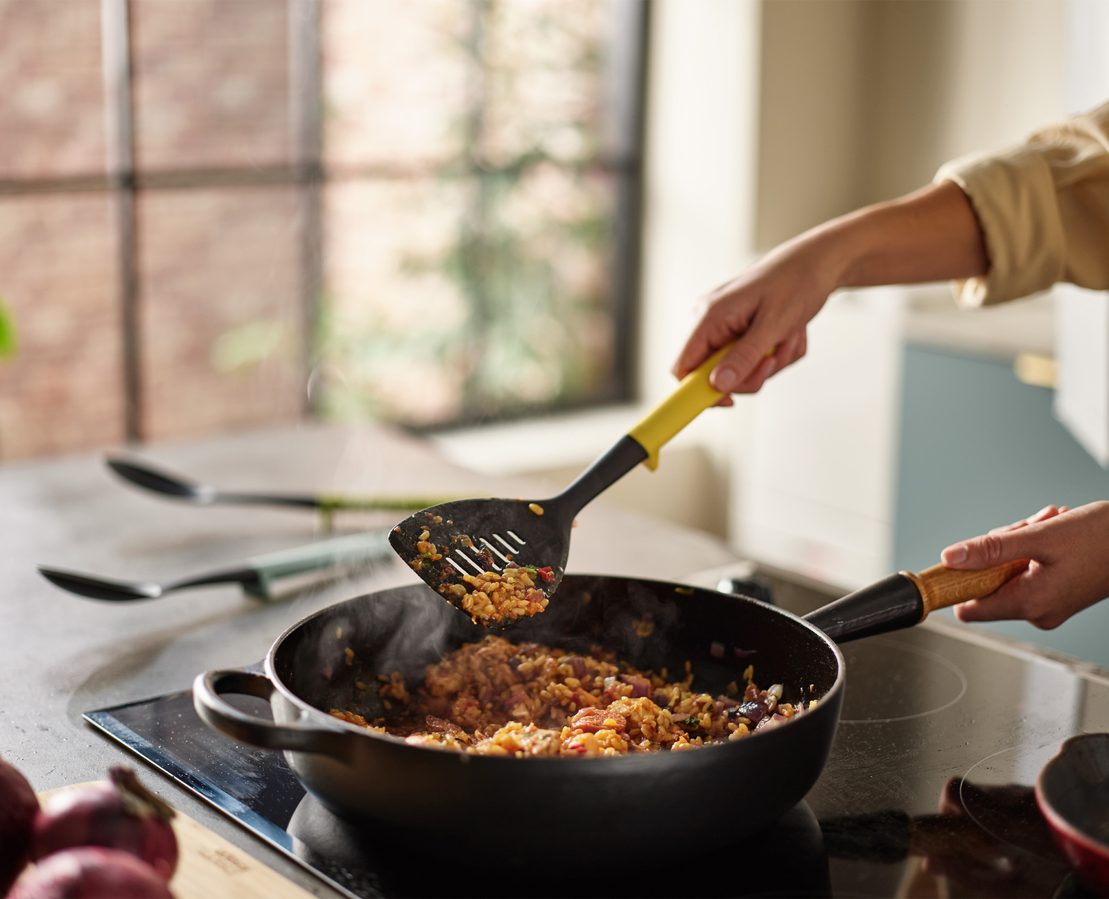 Una persona revuelve comida en una sartén con una espátula. La sartén está en una estufa. El vapor sale de la comida. El fondo muestra la cocina y la ventana.