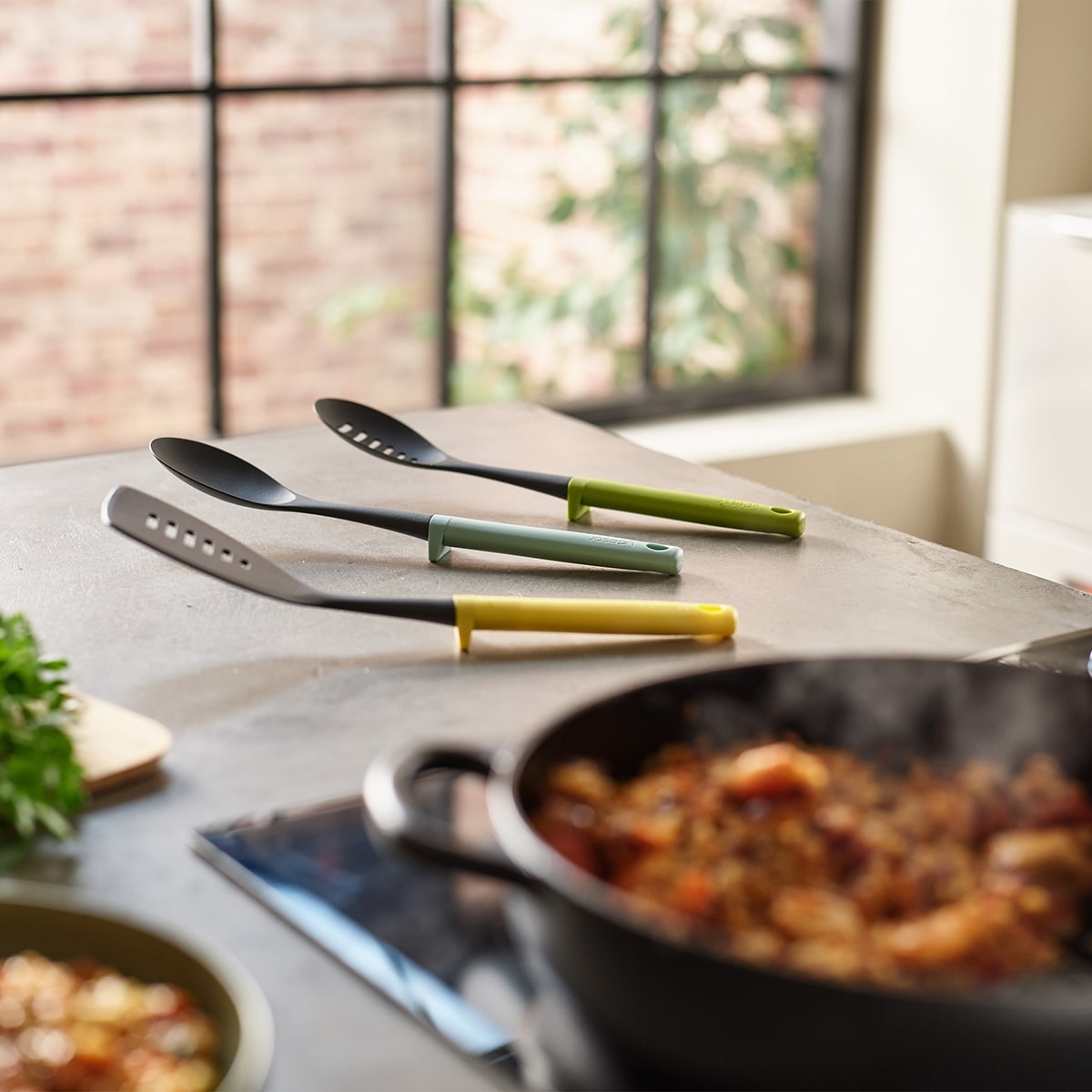 Tres utensilios de cocina en una encimera: una cuchara ranurada, una cuchara sólida y una espátula. Una sartén está en primer plano.