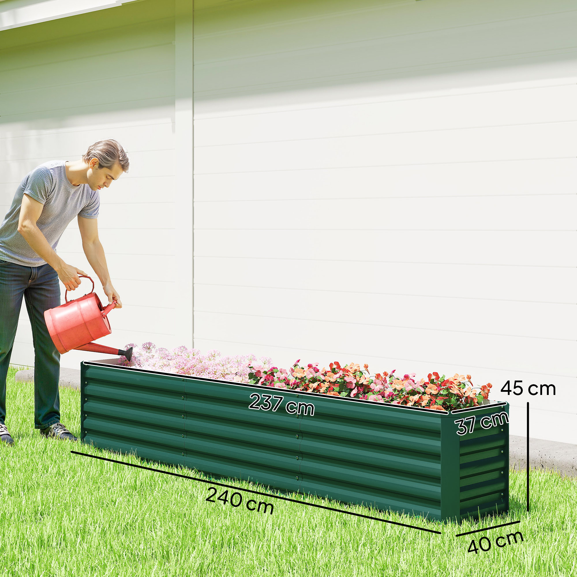 Un uomo annaffia una fioriera rettangolare verde piena di fiori colorati. La fioriera è sull'erba vicino a un muro bianco.