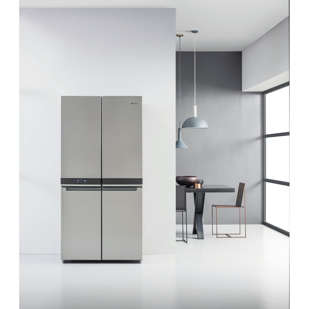 Un moderno refrigerador plateado contra una pared blanca. Un comedor con mesa y sillas en el fondo.