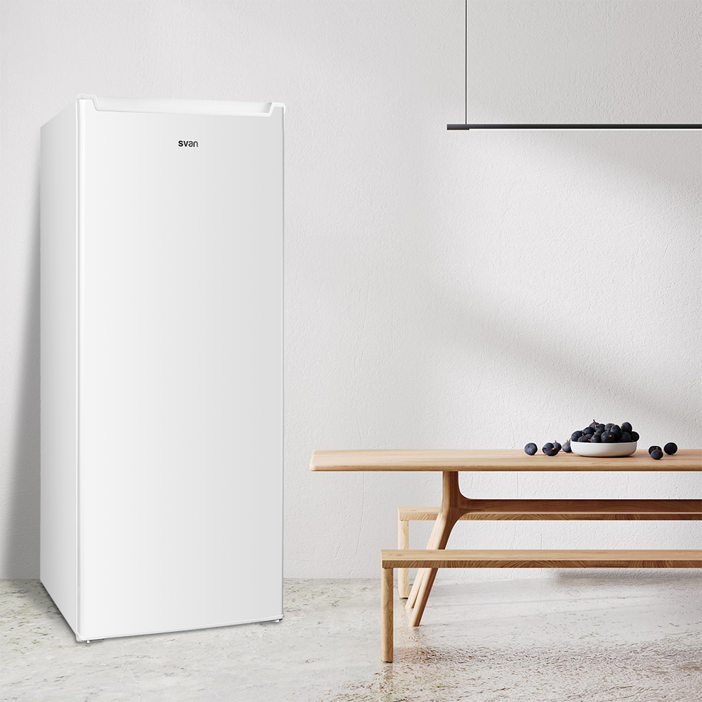 Un refrigerador blanco junto a una mesa de madera con un cuenco de frutas oscuras. Una luz cuelga sobre la mesa.