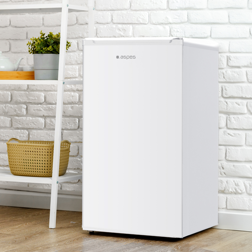 Un mini-refrigerador blanco se encuentra junto a una estantería blanca con una planta y una cesta contra una pared de ladrillos blancos.