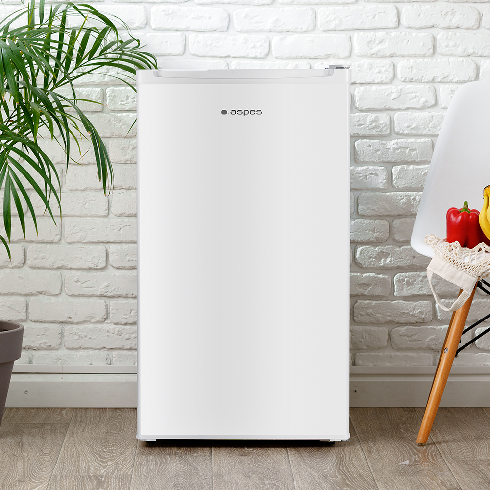 Un refrigerador Aspes blanco frente a una pared de ladrillos blancos. Una planta a la izquierda, una silla con fruta a la derecha.