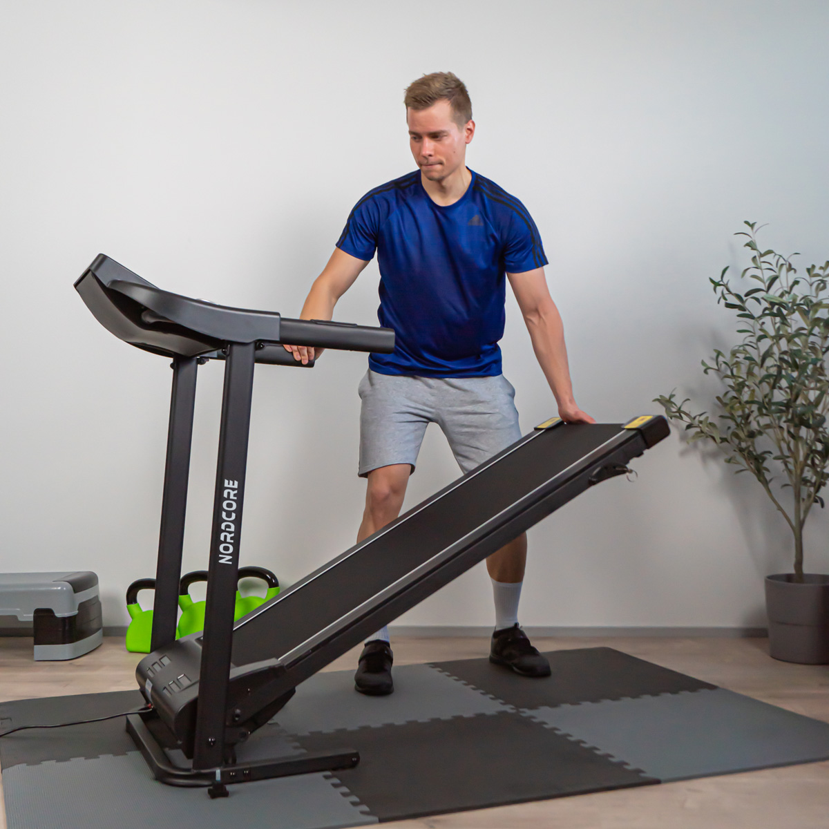Mężczyzna w niebieskiej koszulce i szortach ustawia czarną bieżnię. W tle roślina i inne urządzenia do ćwiczeń.