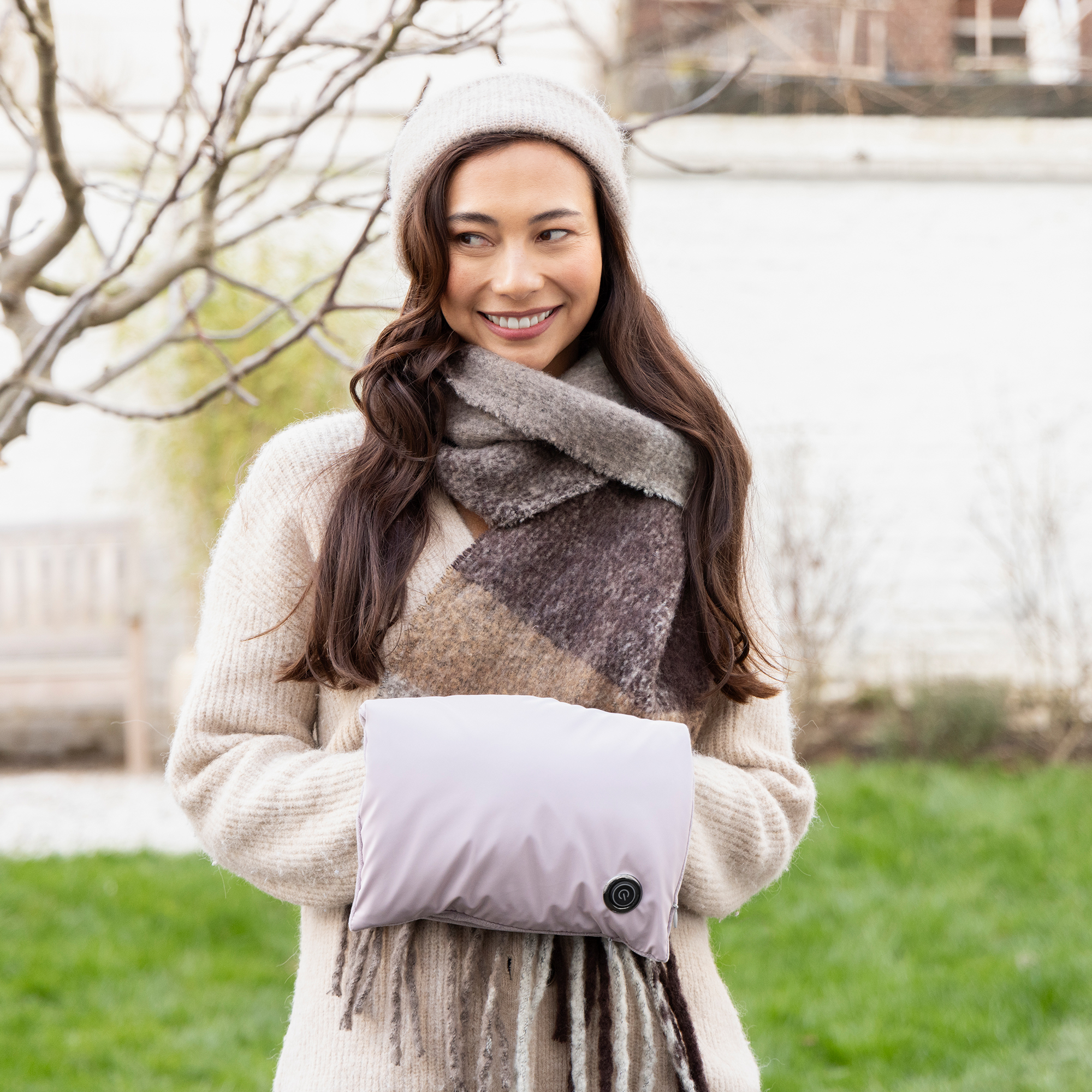 Mujer con gorro y bufanda sosteniendo un calentador de manos lavanda. Sonriendo, de pie sobre la hierba. Fondo con árboles y un edificio.
