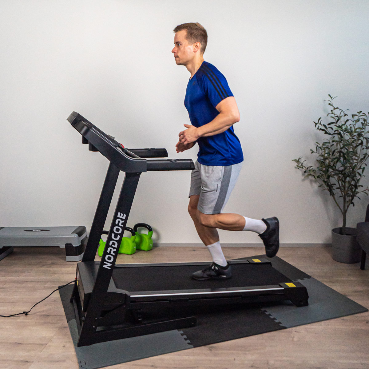 Mężczyzna biega na bieżni. Ma na sobie niebieską koszulę, szare szorty i czarne buty. Tło zawiera sprzęt do ćwiczeń i roślinę.