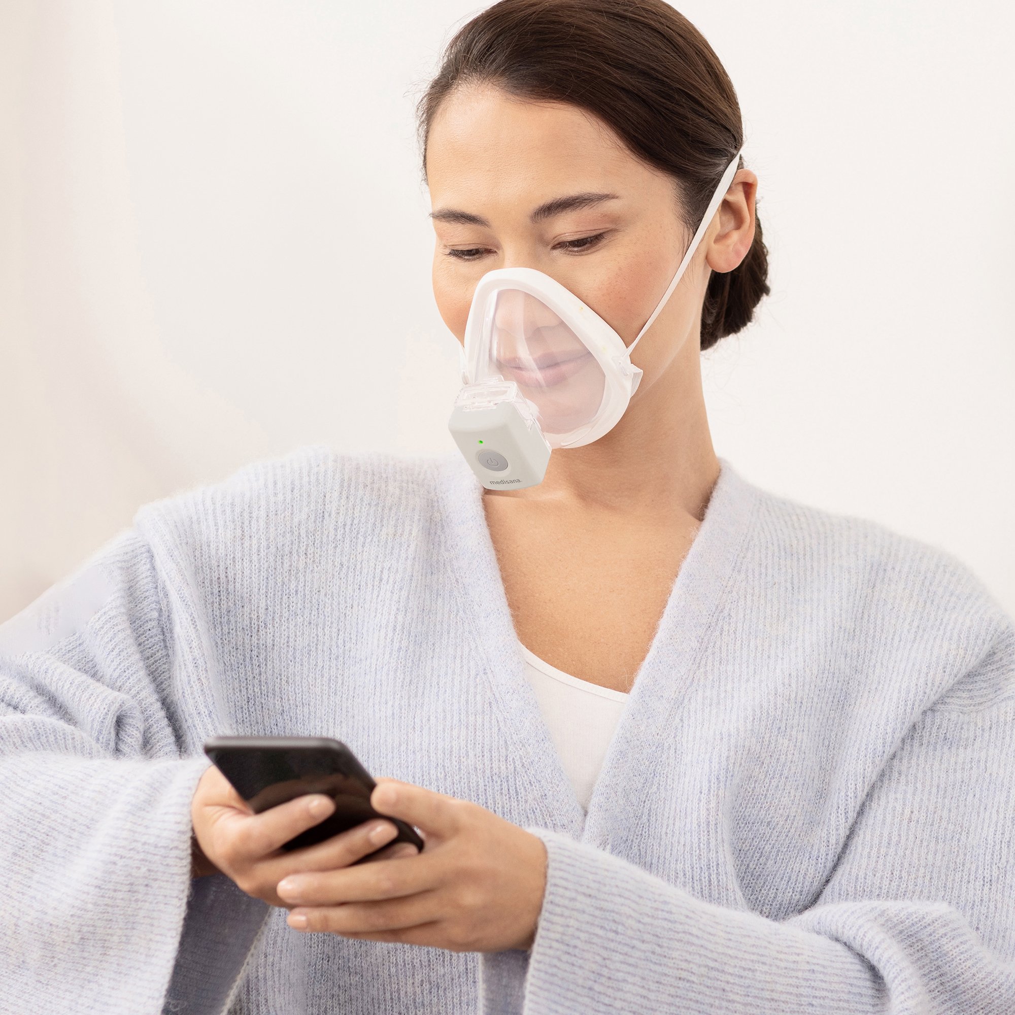 Woman wearing a face mask and looking at a smartphone. She is wearing a light blue sweater.