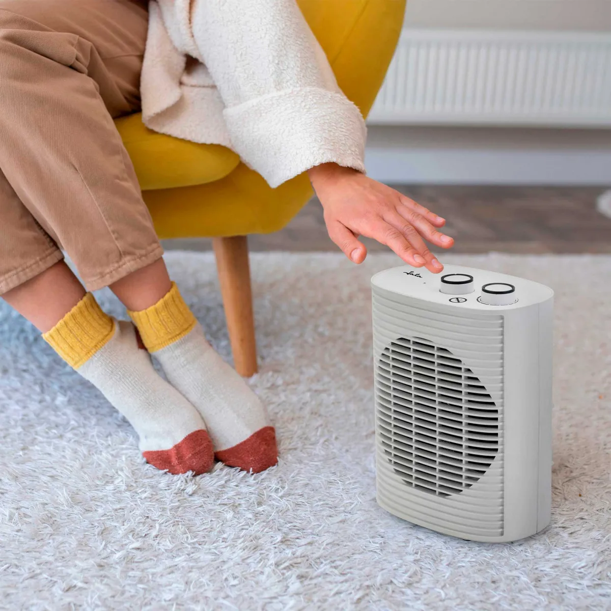 Eine Person berührt eine weiße Heizung. Sie sitzt auf einem gelben Stuhl. Die Person trägt Socken und eine weiße Jacke.