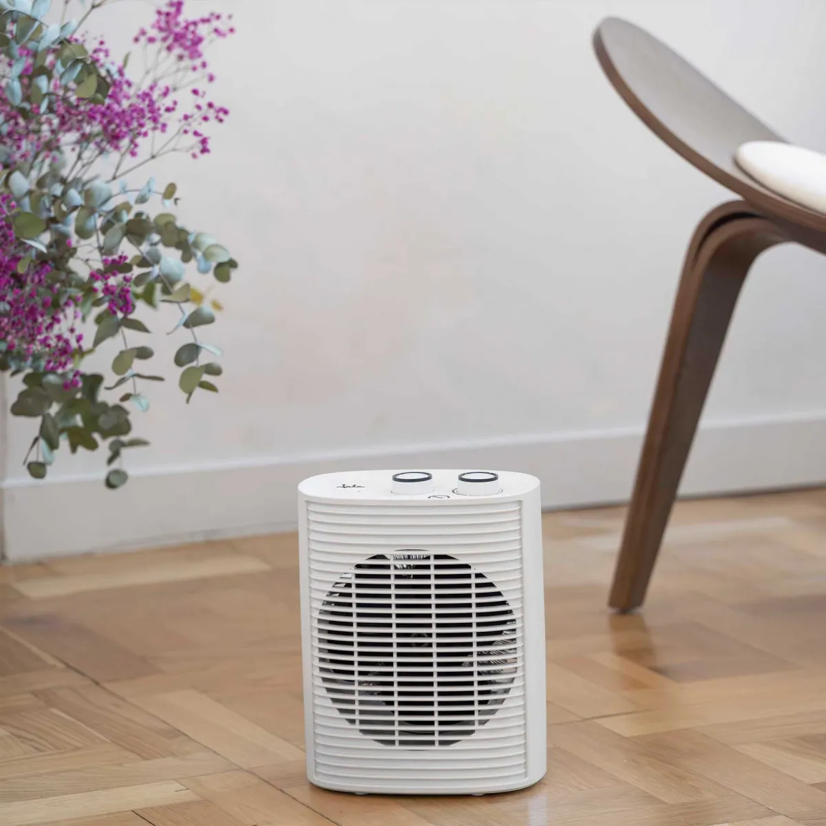 Ein weißer Elektroheizer steht auf einem Holzboden in der Nähe eines Holzstuhls und Blumen.