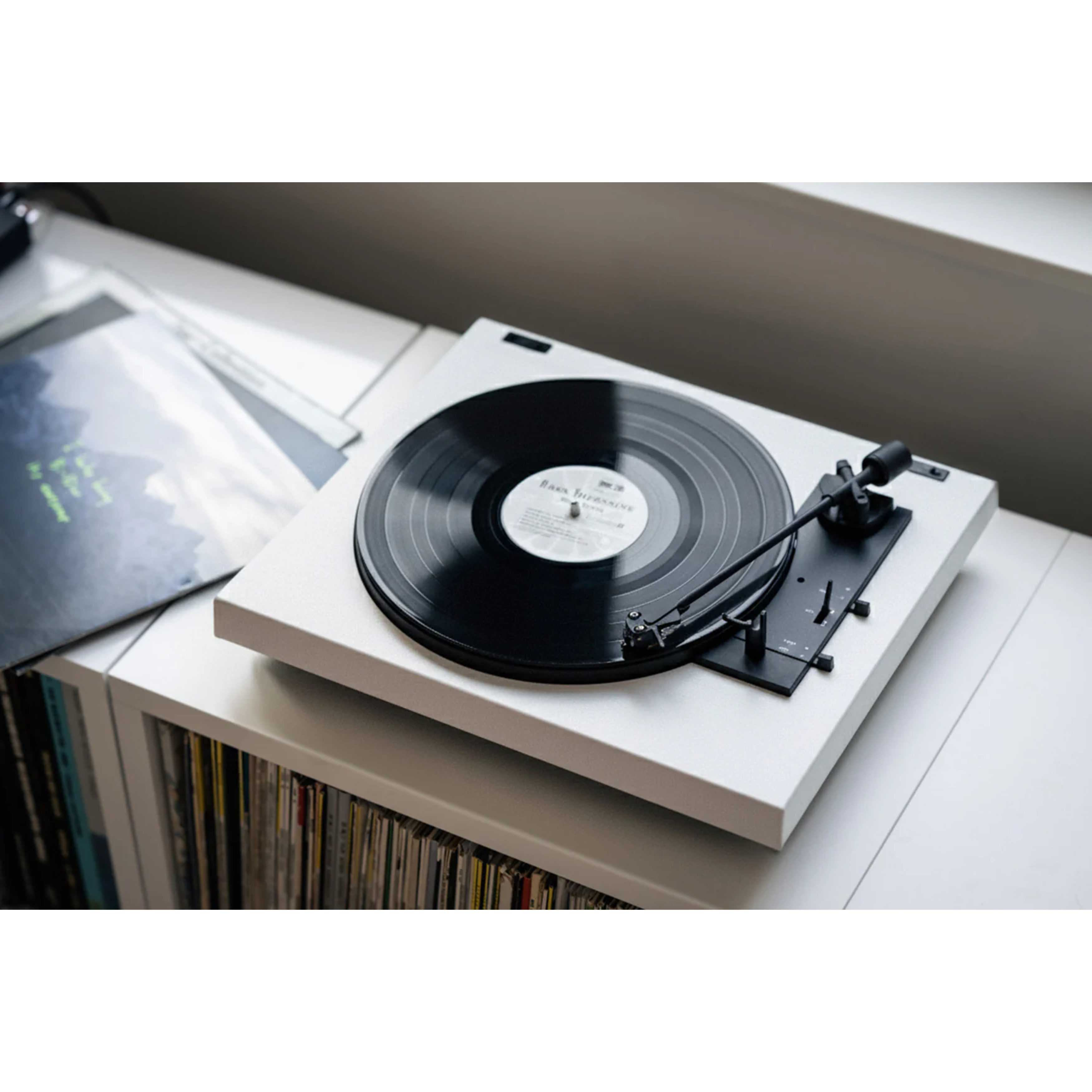 Een witte platenspeler met een zwarte vinylplaat. Andere platen staan op een plank eronder. Een raam staat op de achtergrond.