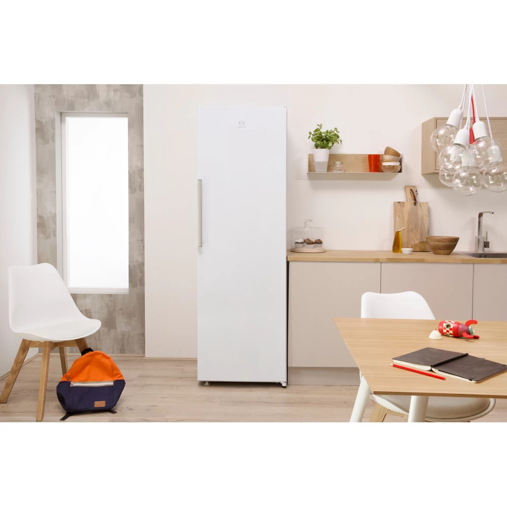 Un refrigerador blanco está en una cocina. Hay una mesa con cuadernos y una silla. Una mochila está en el suelo.