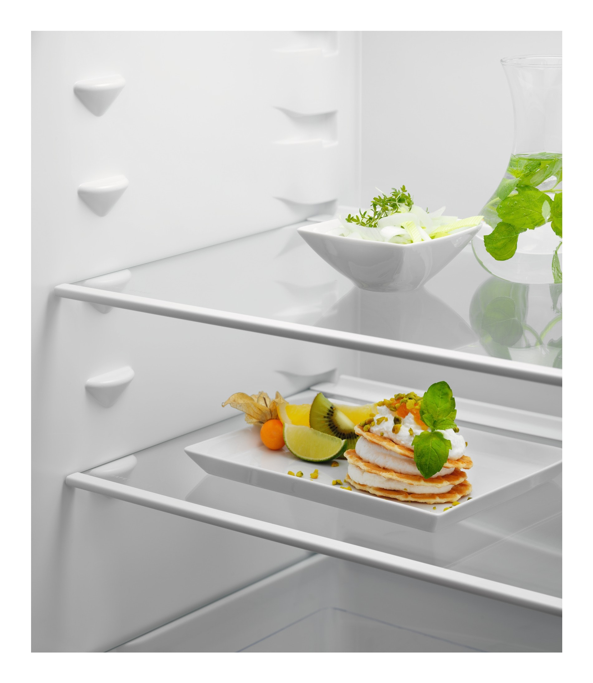 Wnętrze lodówki z jedzeniem na półkach: deser, owoce i zioła. Na górnej półce szklanka wody z infuzją.