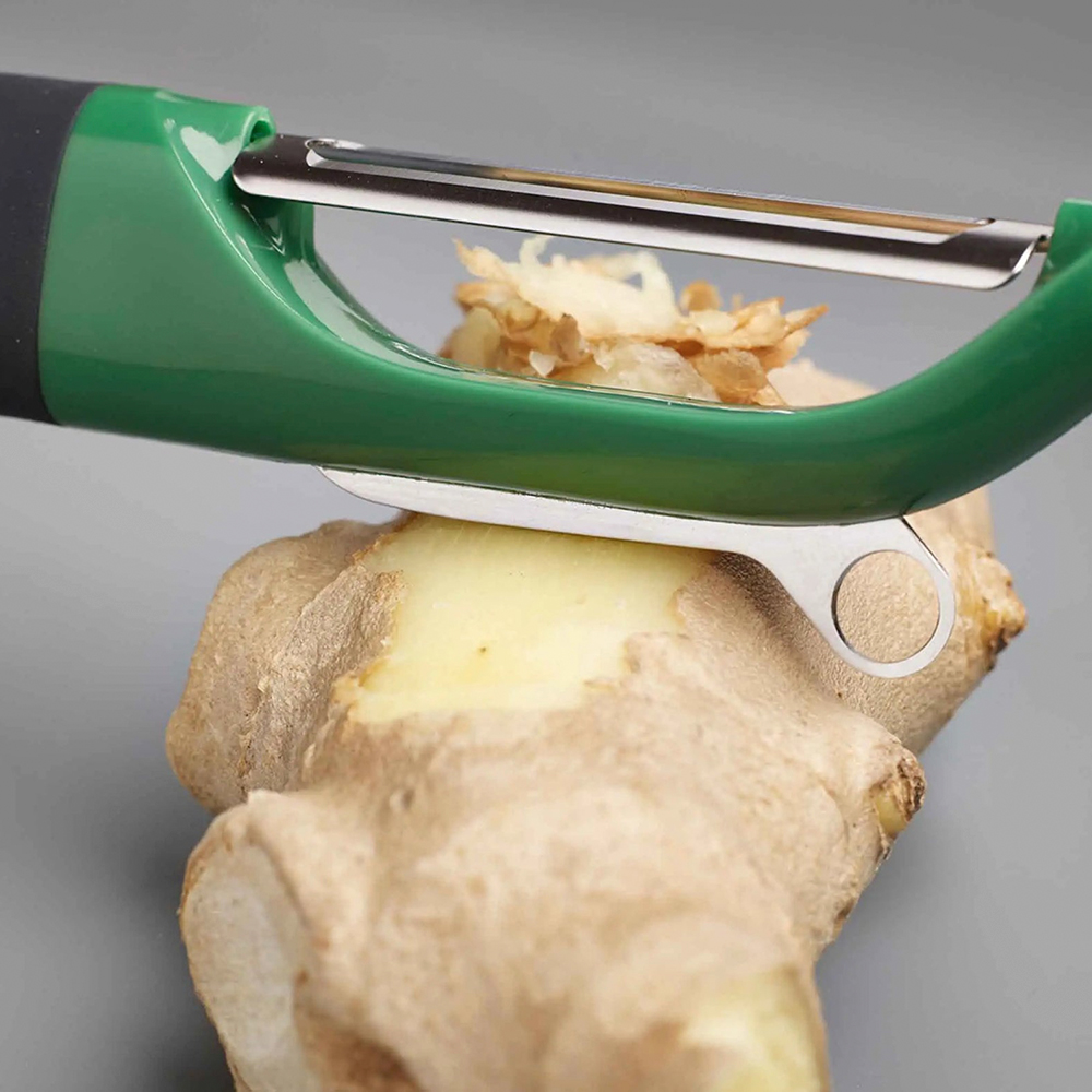 A green peeler removing the skin from a ginger root. Ginger shavings are visible. The background is a plain gray color.
