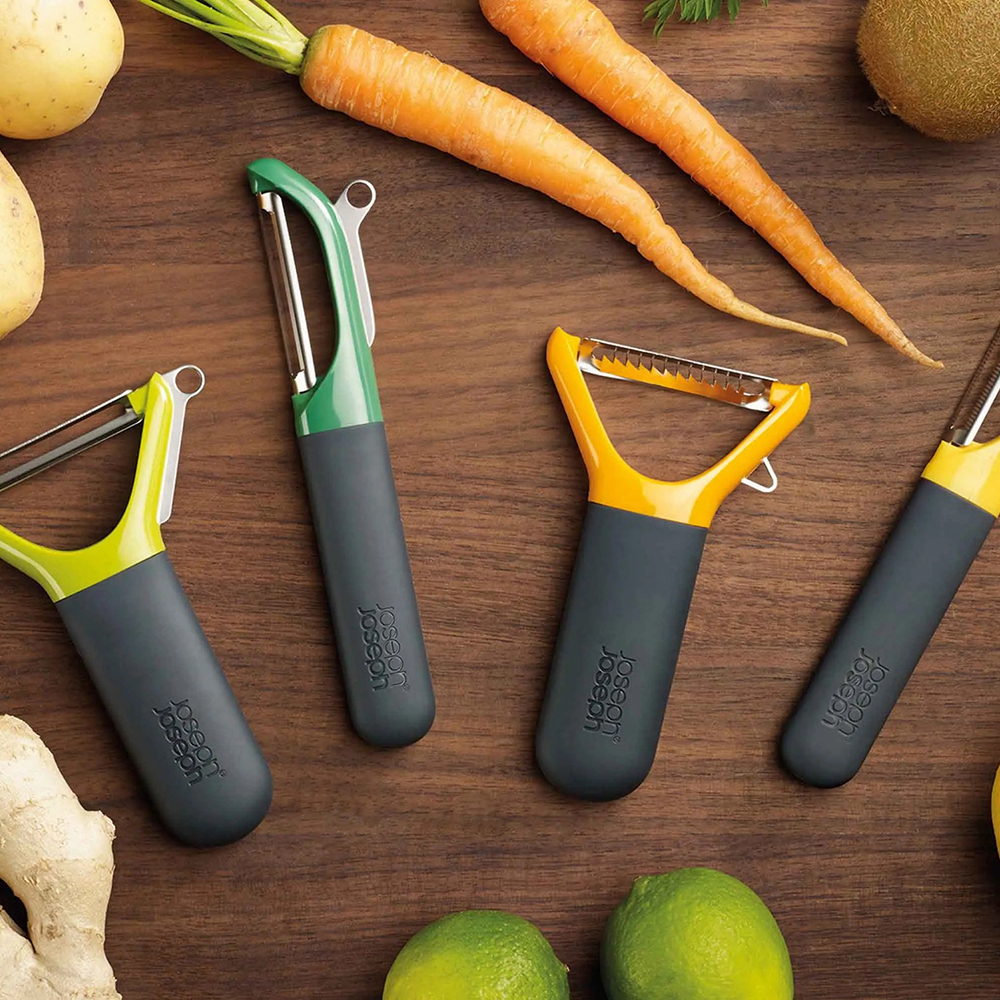 Four colorful peelers on a wooden surface with carrots, potatoes, limes, and ginger.