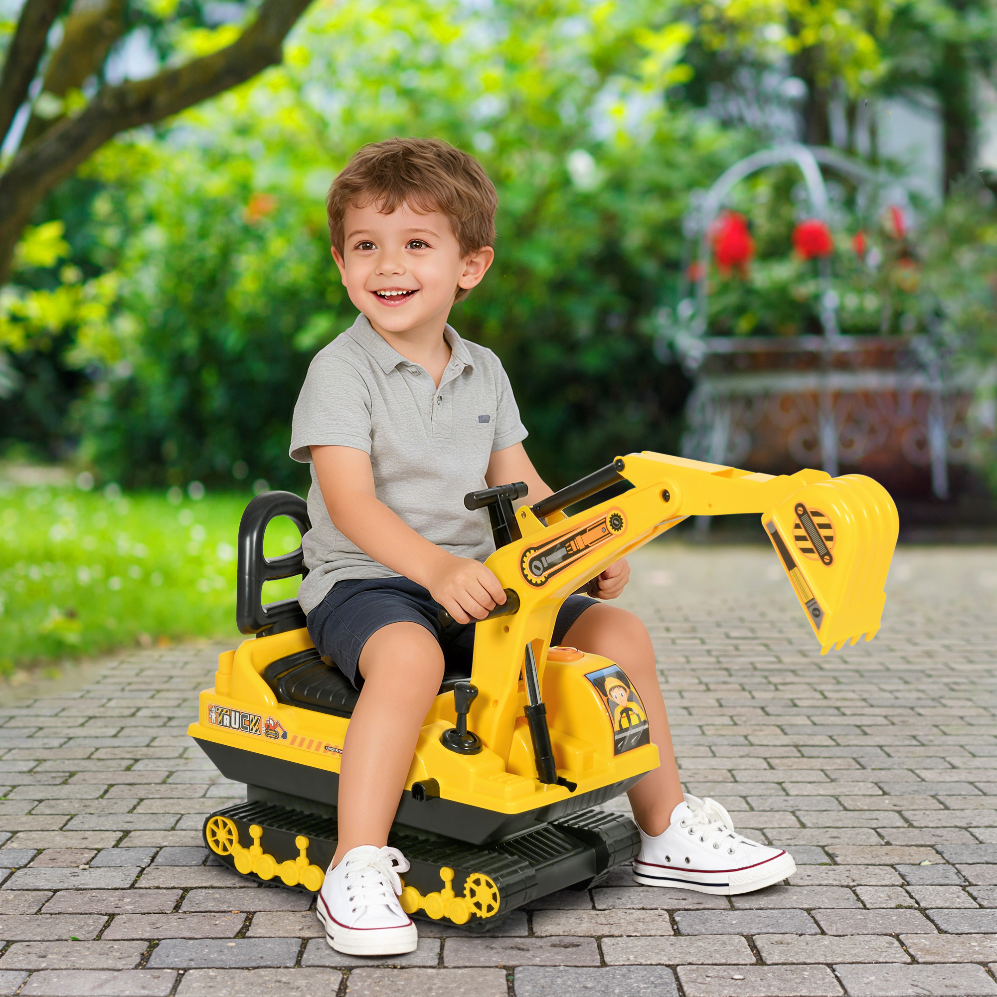 Un niño sonriente está sentado en una excavadora de juguete amarilla. Lleva una camisa gris, pantalones cortos y zapatillas blancas. El fondo es un jardín.