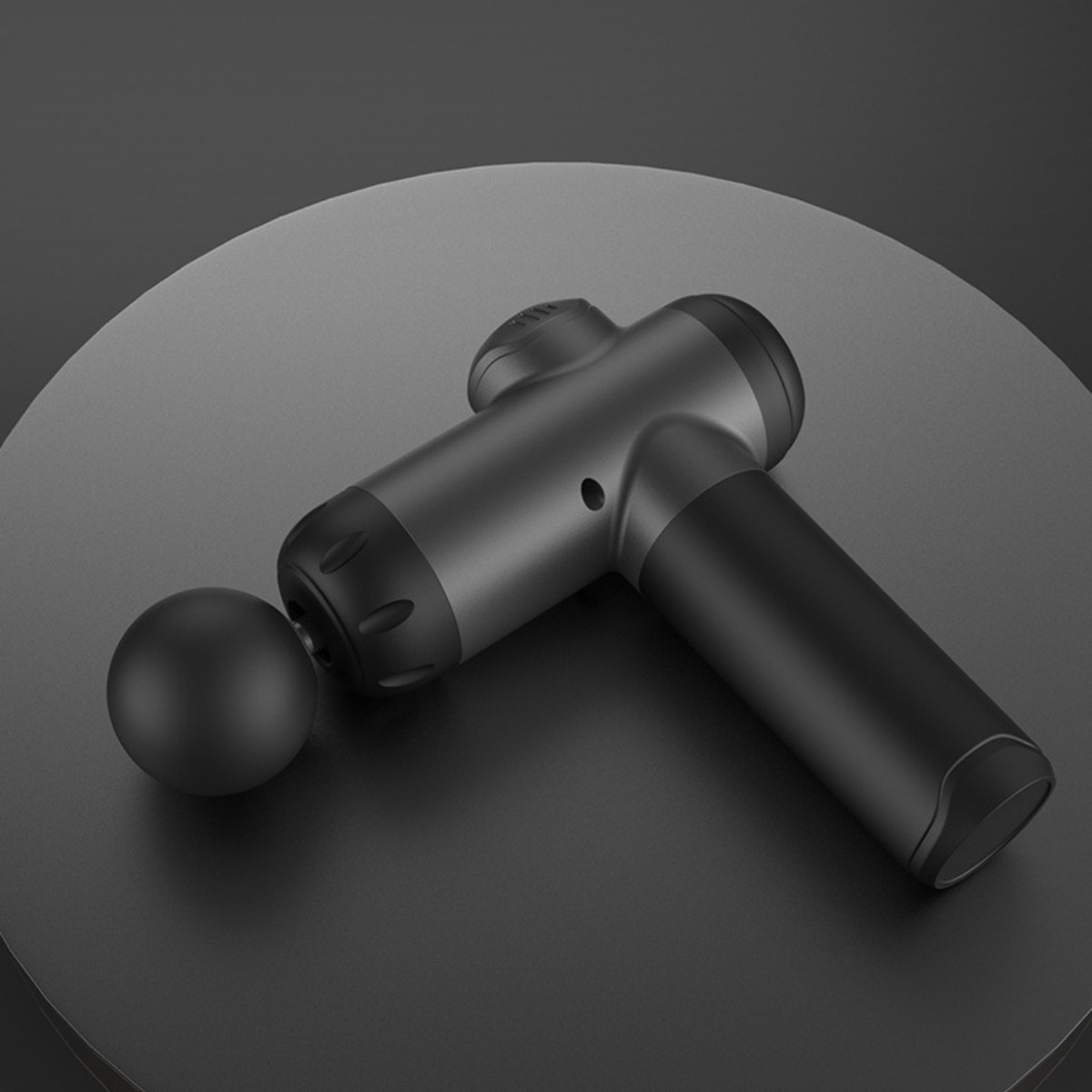 Eine schwarze Massagepistole und ein schwarzer Ball liegen auf einer schwarzen, kreisförmigen Oberfläche. Die Pistole ist angewinkelt, der Ball links.