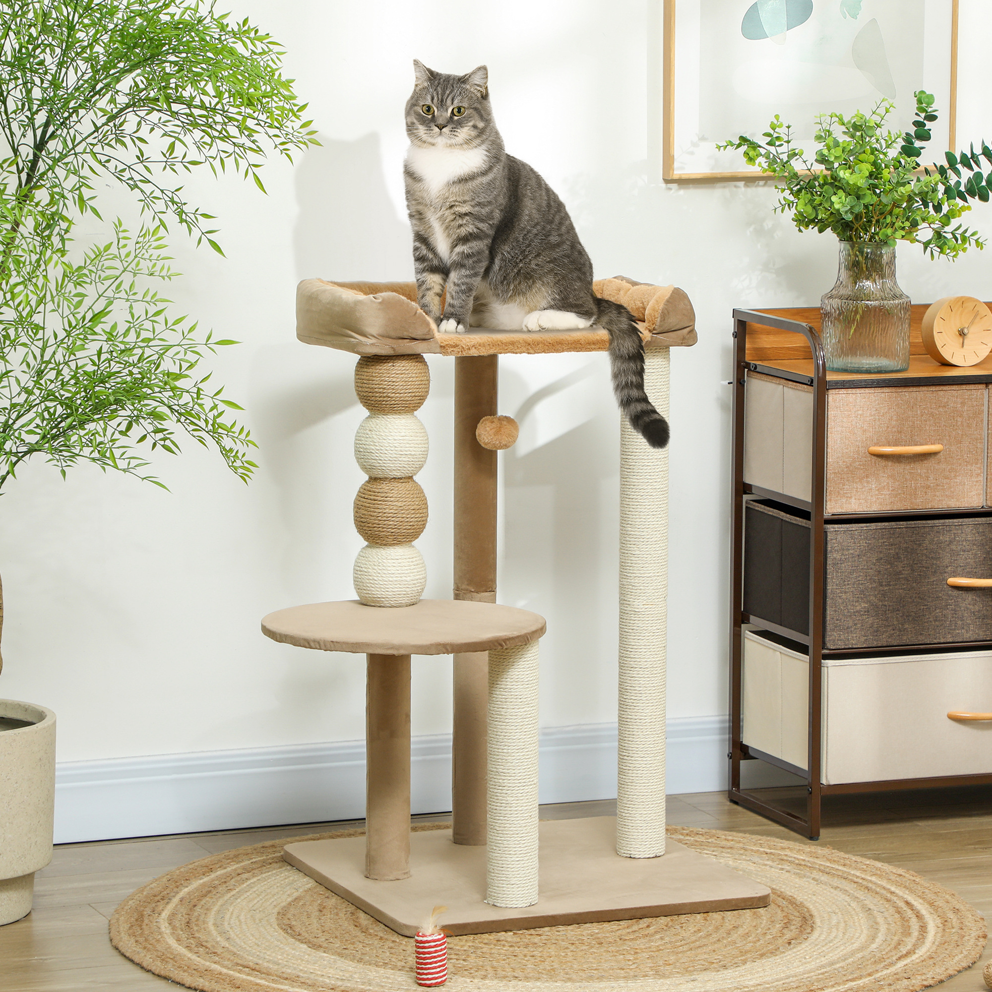 Un gato gris está sentado en un árbol para gatos. El árbol tiene múltiples niveles y postes para rascar. El fondo tiene plantas y muebles.